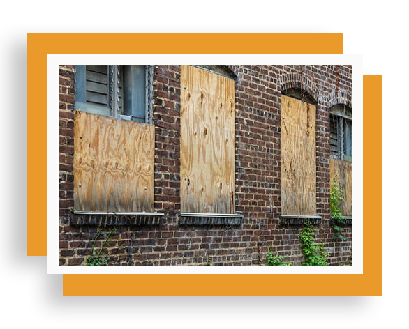 A brick building with boarded up windows and plants growing on the side.